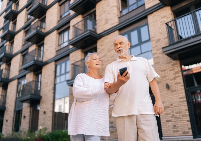 Shooting from below of cheerful elderly couple standing embracing on sunny day, using looking smartphone in front of modern brick apartment complex, exuding togetherness and love.